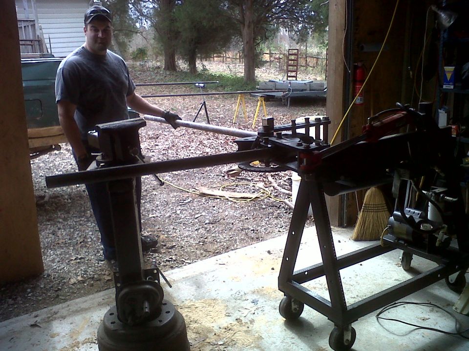 Man operating a pipe bender mounted on a stand, with a vise on another stand. Garage setting