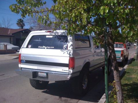 1996 Ford C-350 (Centurion Conversions) - White Ford Bronco parked curbside, partially obscured by a tree. Another truck is visible behind it.