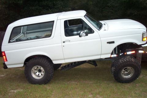 1995 Ford Bronco - Passenger-side profile of a white lifted full-size Ford Bronco with BRONCO XLT fender badging, riding on tall aggressive mud tires with mac…