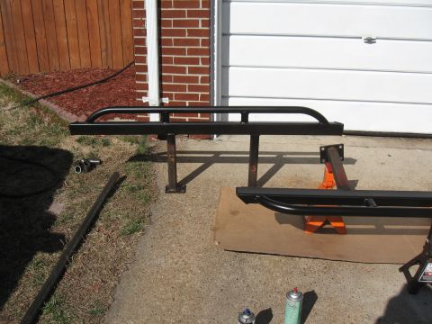 1994 Ford Bronco - Two black metal rock sliders are displayed on jack stands in front of a garage.
