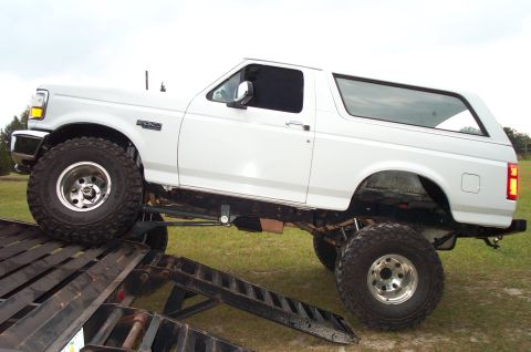 1995 Ford Bronco - Driver-side view of a heavily lifted white full-size Ford Bronco with a matching hardtop, being driven off a black steel trailer ramp onto…