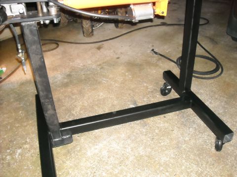 1979 Ford F-150 - Close-up of a metal engine hoist stand with wheels, with a portion of an orange machine in the background.