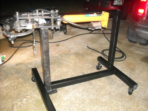 1979 Ford F-150 - A hydraulic tube bender is set up on a rolling stand, with parts of vehicles visible in the background.