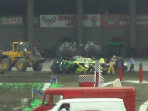 1986 Ford Bronco - A monster truck has overturned during a Monster Jam event, with a yellow loader and several people attending the scene.
