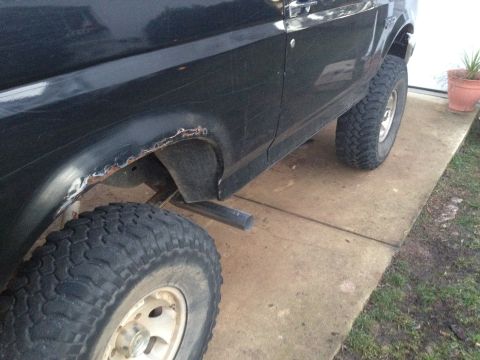 1995 Ford Bronco - The side of a black SUV shows significant rust near the rear wheel arch and is equipped with large off-road tires.