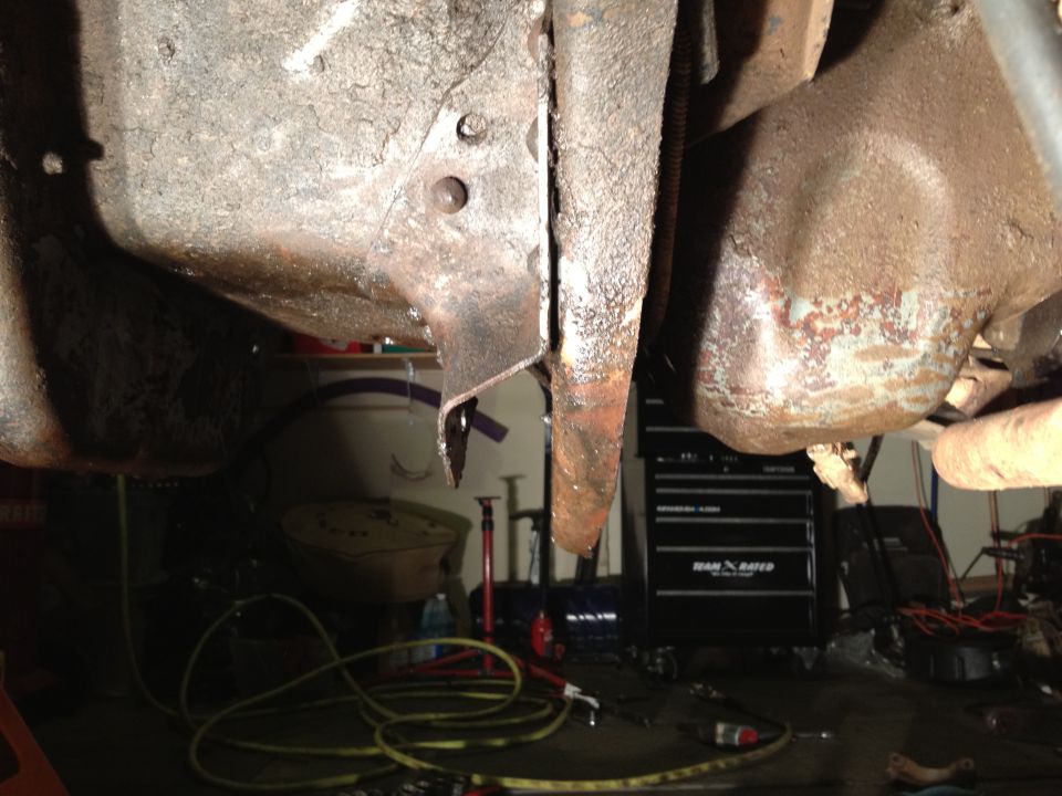 Underside of a vehicle showing rusty frame components and suspension parts. A toolbox is visible in the background