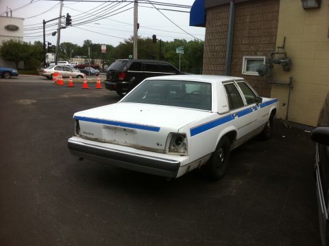1991 Ford LTD Crown Victoria - A white sedan with a blue stripe is parked outside a building, showing damage to the rear.