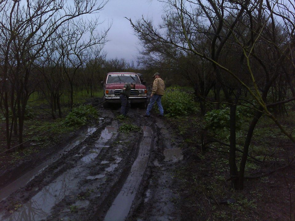 Red Ford Bronco stuck in mud on a dirt road, front view. Two people are near the vehicle