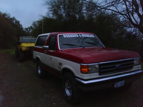 1989 Ford Bronco - A red and white 1989 Ford Bronco is parked outdoors with a yellow Jeep in the background.