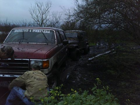 1989 Ford Bronco - A 1989 Ford Bronco and another SUV are stuck in the mud, with people working on the front vehicle.
