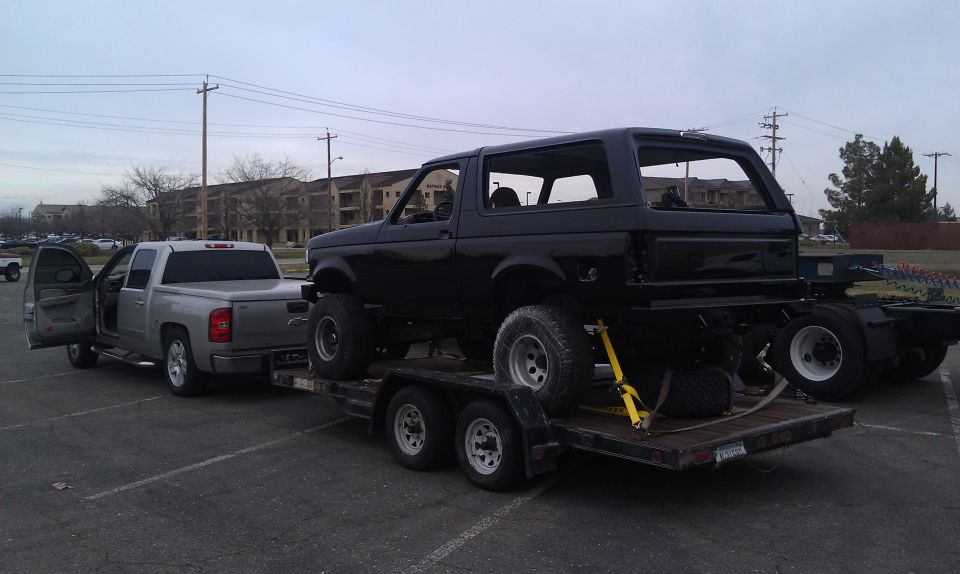 Black Ford Bronco on a dual-axle trailer, towed by a silver pickup truck