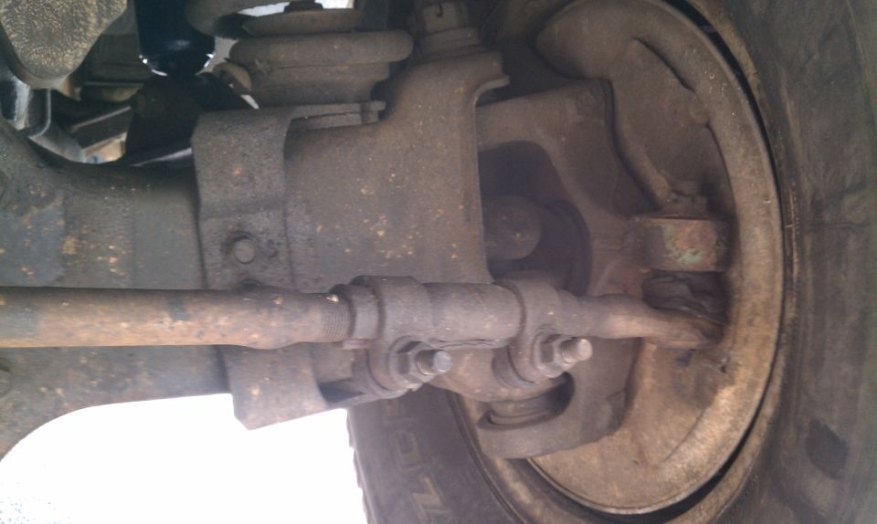 Underside view of rusty front tie rod, steering knuckle and drum brake behind tire on 1984 Ford Bronco