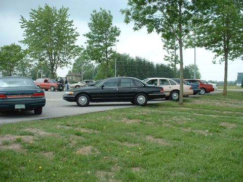 2000 Ford Crown Victoria - Several cars are parked in a lot with grass and trees surrounding them.