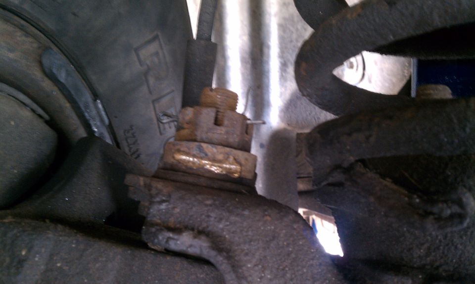 Underside close-up of rusty castle nut and cotter pin on a suspension joint near coil spring and tire