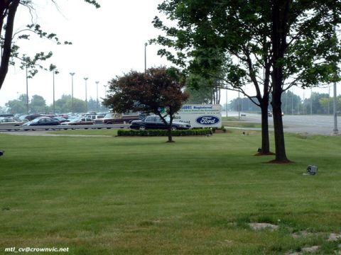 2000 Ford Crown Victoria - A black sedan is parked in front of a Ford dealership sign, with a large parking lot full of vehicles in the background.