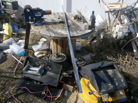 1979 Ford Bronco - A cluttered outdoor scene with various tools and equipment, including a vise on a wooden spool, a winch, and parts of a lawnmower. A metal…