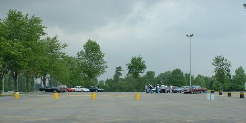 2000 Ford Crown Victoria - A group of people gather around several cars parked in an outdoor lot on an overcast day.