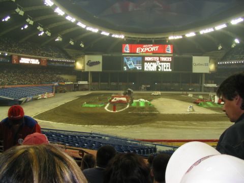 1986 Ford Bronco - A monster truck event is underway in a large indoor stadium with a dirt track and multiple ramps.