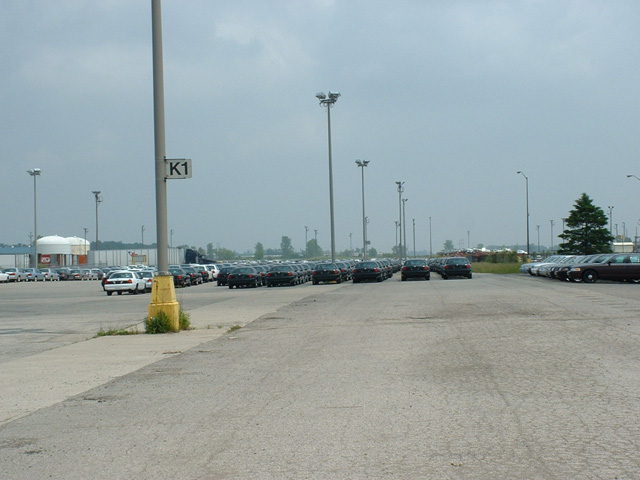 Rows of parked sedans in a large plant lot beside a light pole marked K1