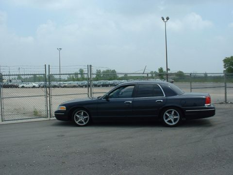 2000 Ford Crown Victoria - A dark blue 2000 Ford Crown Victoria is parked in an outdoor lot with a chain-link fence and other vehicles in the background.
