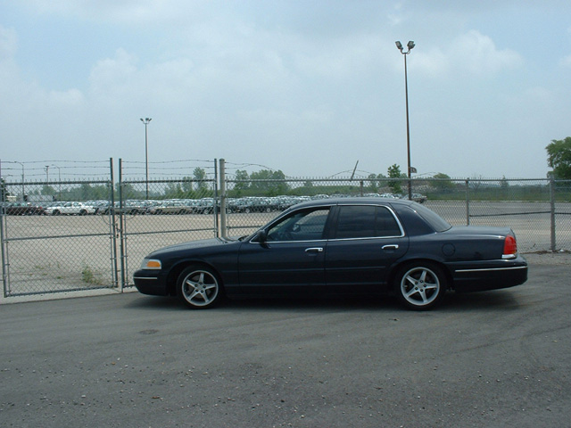 Lowered dark blue Ford Crown Victoria in side profile beside a chain-link fence and storage lot