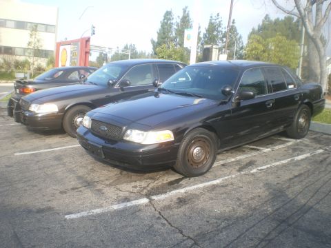 2004 Ford Crown Victoria - Two Ford Crown Victoria police cars are parked in a lot. The front car is black with steel wheels.