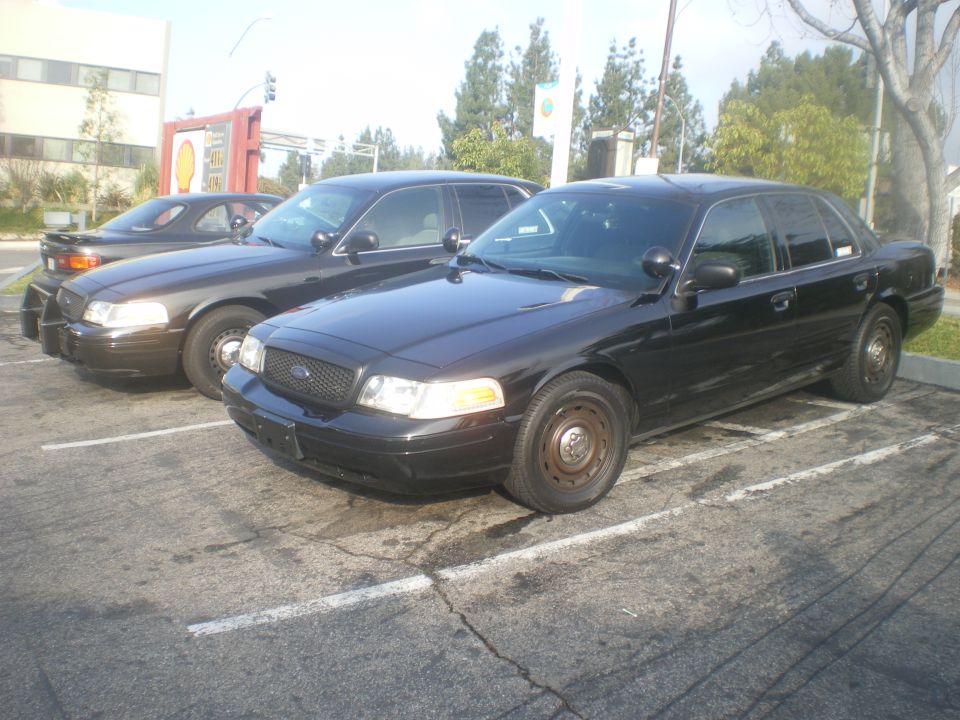 Two black Ford Crown Victoria police cars parked side-by-side, one in foreground