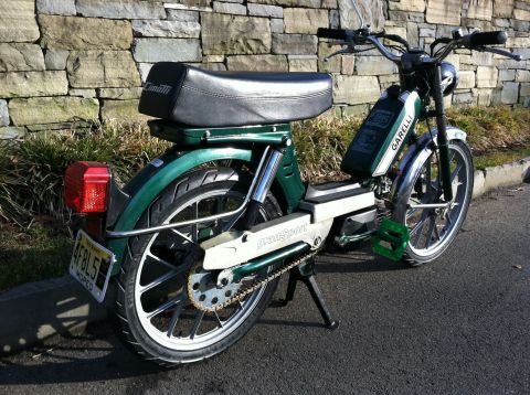 1981 Garelli Rally Sport - A green moped is parked on the street in front of a stone wall. The moped has a black seat and green pedals.
