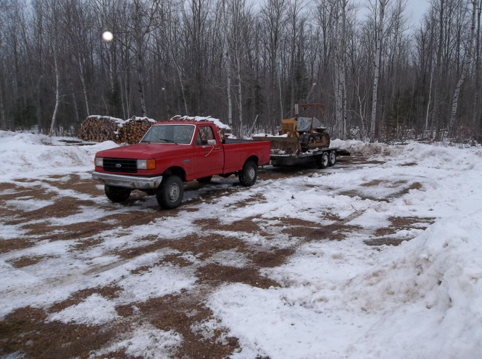Red 1987 Ford F-250 pickup truck towing a bulldozer on a trailer in a snowy, wooded area