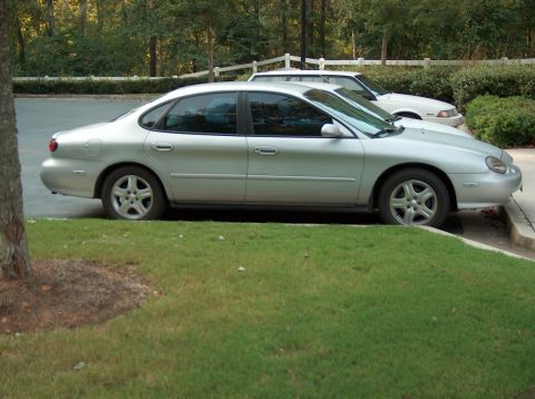 1998 Ford Taurus - A silver sedan is parked on a grassy area next to a curb, with another white vehicle visible behind it.