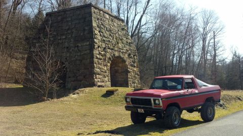 1979 Ford Bronco - A red pickup truck with a lifted suspension is parked beside an old stone building in a wooded area.