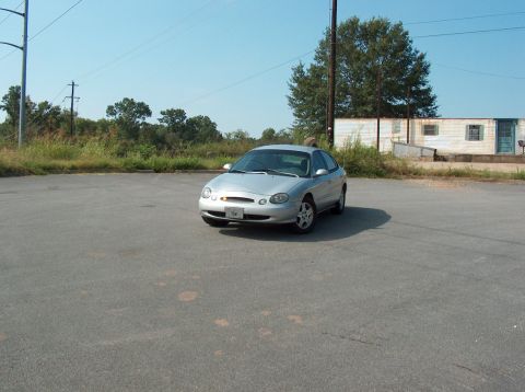 1998 Ford Taurus - A silver sedan is parked in an empty lot with a building and trees in the background under a clear blue sky.