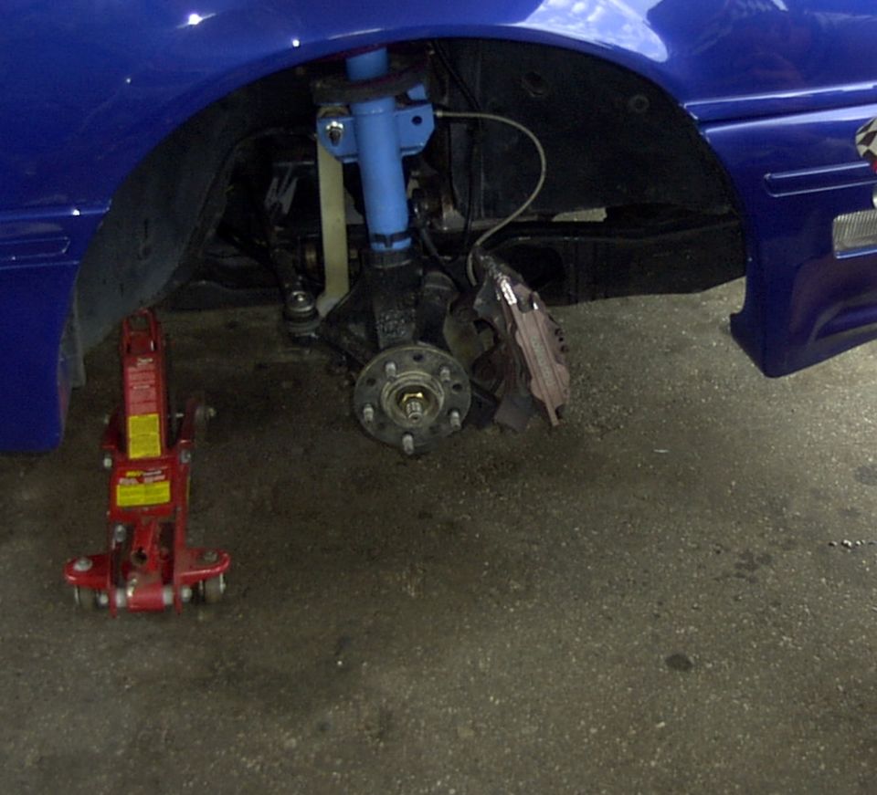 Blue car's front wheel hub, brake caliper, and suspension components are visible with a red hydraulic jack