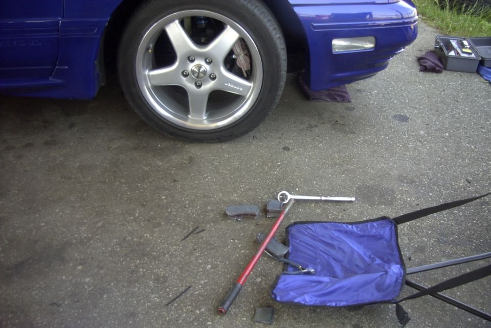 Blue car's front wheel and fender with tools on the ground, including a lug wrench and a blue bag
