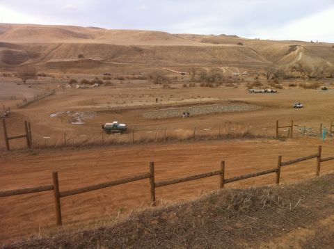 1989 Ford Bronco - A light-colored pickup truck is seen driving through a dusty, rural area with sparse vegetation and rolling hills.