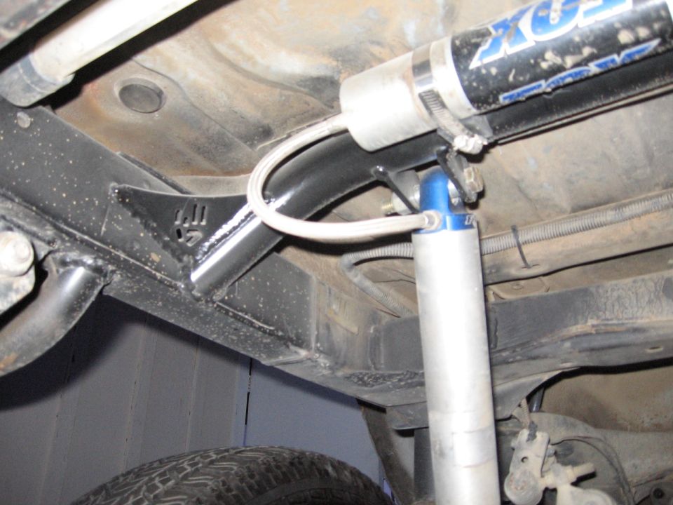Vehicle undercarriage with black frame, silver shock absorber with blue top, and a black hose. Text 'SUPERMOTORS.NET' visible