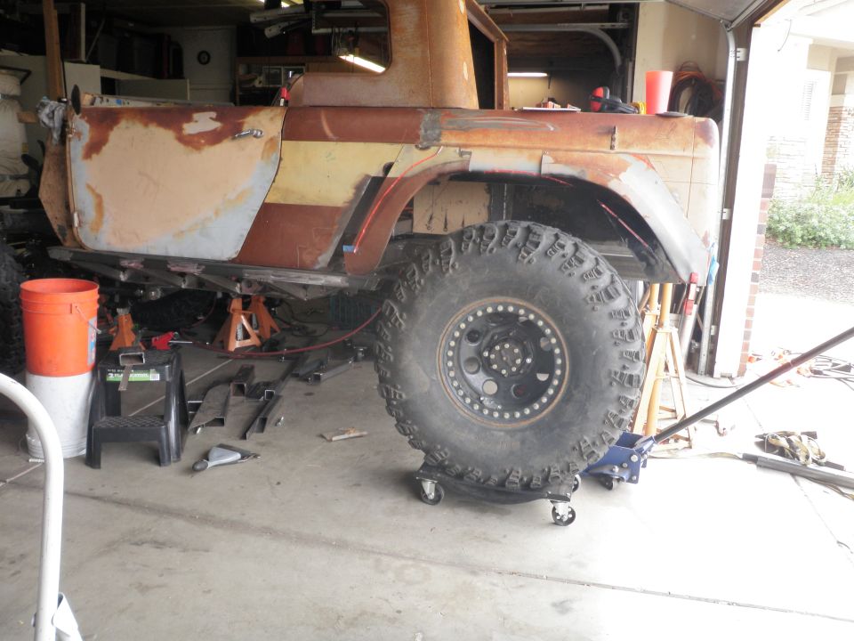 Rusted vehicle body on jack stands, large off-road tire, metal fabrication parts scattered around