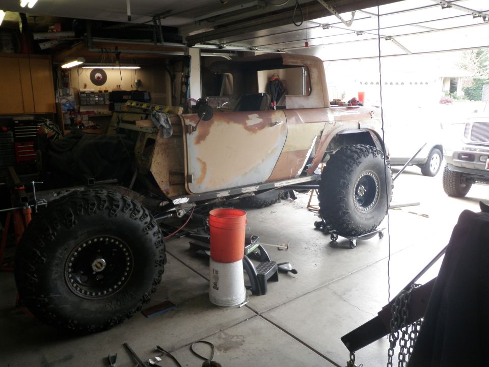 Custom off-road vehicle body in garage, undergoing modifications. Large tires are mounted. Another SUV is visible in the background