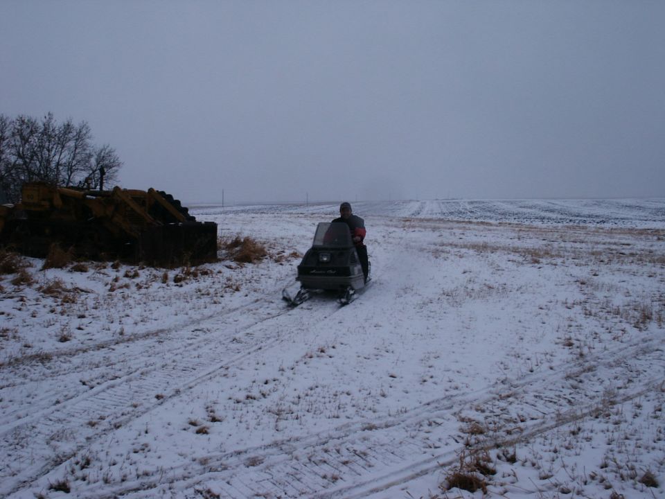Snowmobile with rider on a snowy track in a field, with a pile of debris to the left