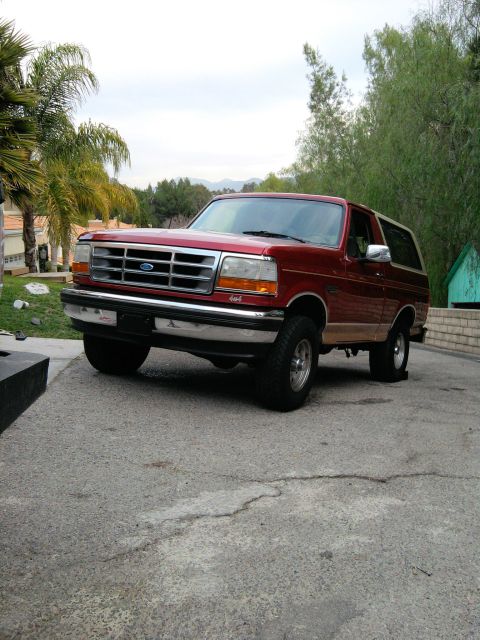 1994 Ford Bronco - This 1994 Ford Bronco is shown in a three-quarter front view, parked on a paved surface with trees and a house in the background.