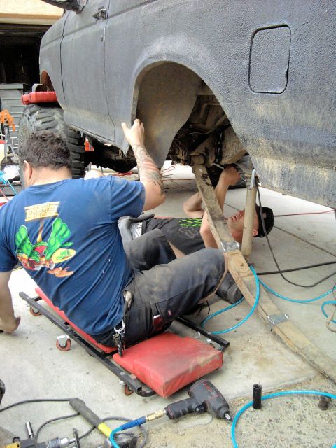 1994 Ford Bronco - Two people work under the passenger-side rear quarter of a lifted SUV finished in flat black bedliner-style coating, one seated on a red ro…