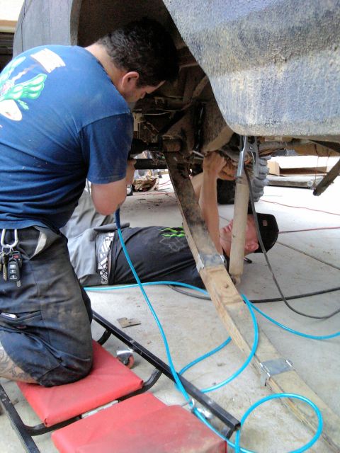 1994 Ford Bronco - Two people work under the raised rear of a truck in a driveway: one kneels on a red creeper pad while the other lies on the concrete beneat…