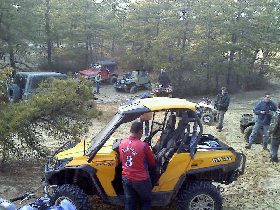 Yellow Can-Am side-by-side with people around it, plus several Jeeps and ATVs in a wooded area
