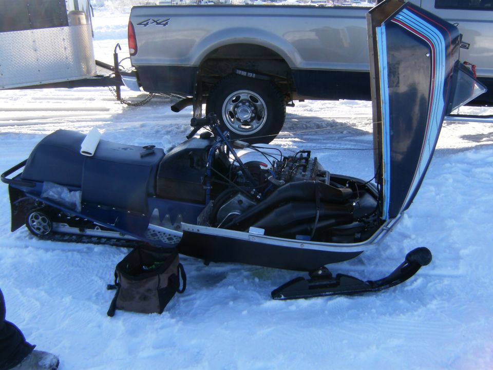 Snowmobile with hood open showing the engine, parked in the snow next to a pickup truck and trailer
