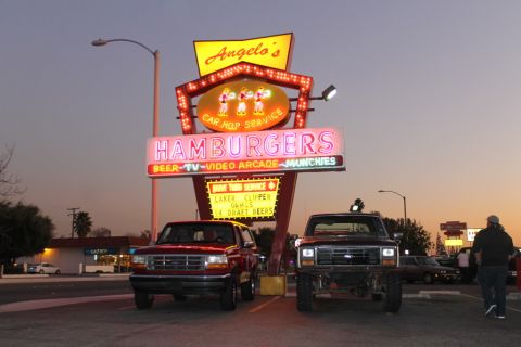1994 Ford Bronco - Two Ford trucks parked nose-out at dusk beneath the tall neon "Angelo's Car Hop Service / HAMBURGERS" drive-in sign: a red 1992-96 Bronco o…