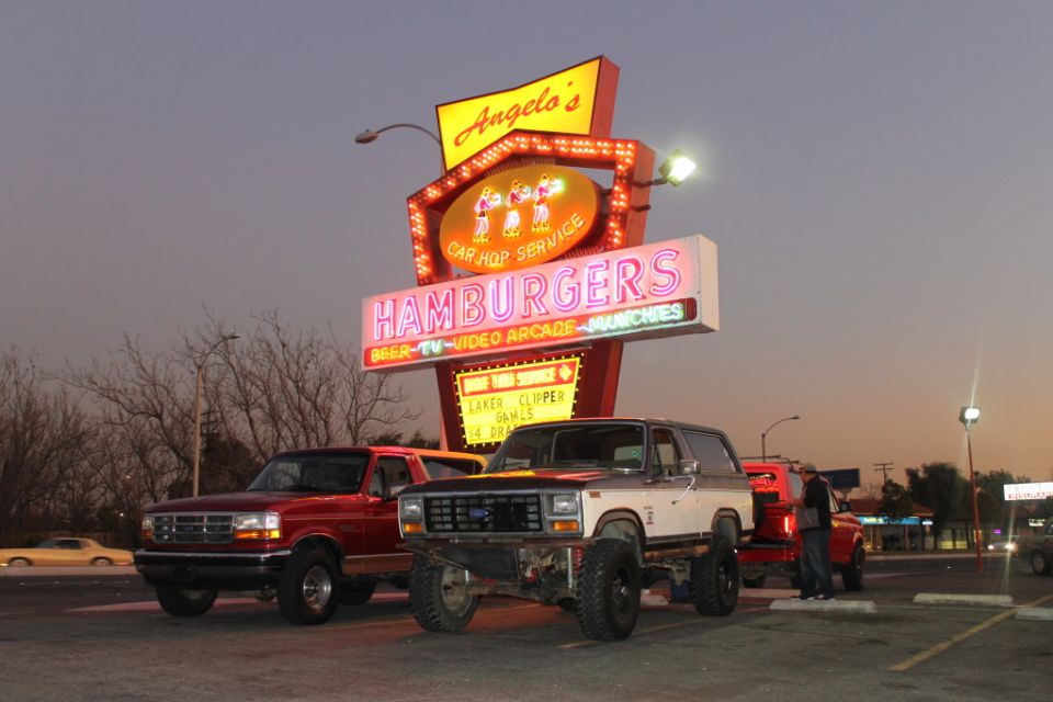Red 1990s Ford Bronco next to lifted red and white 80s Bronco under Angelo's neon sign at dusk