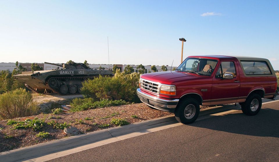 Red Ford Bronco parked by a curb with a military tracked vehicle marked Oakley 453 behind it