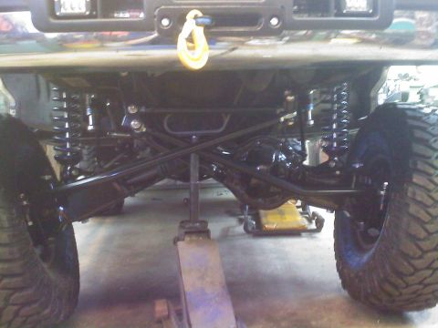 1996 Ford Bronco - Front undercarriage of a lifted 4x4 raised on a jack, showing a solid front axle, dual coilover shocks, tubular control arms and large mud-…