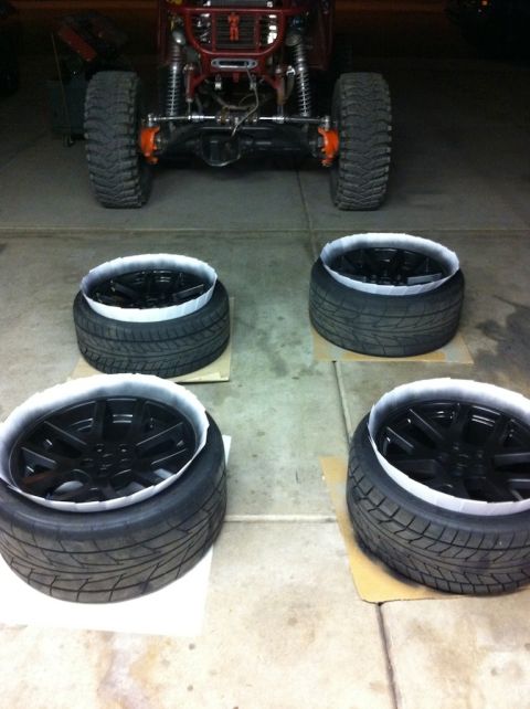 2010 Dodge Challenger - Four black rims are placed on tires, ready for installation, with a red off-road vehicle visible in the background.