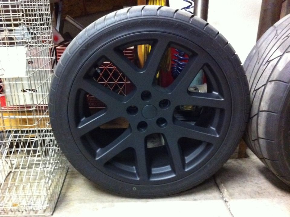 Flat gunmetal-painted five-spoke alloy wheel with low-profile tire standing in a garage